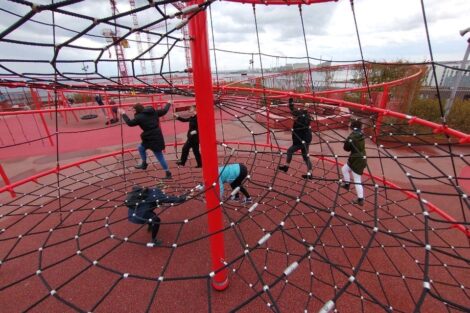 Six students are climbing in a red jungle gym. They are hanging onto the rope netting.