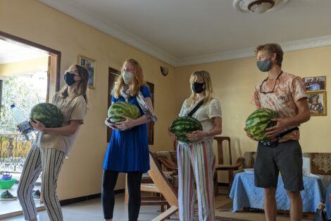 Four students are standing in a living room with yellow walls. They're wearing masks and holding watermelons.
