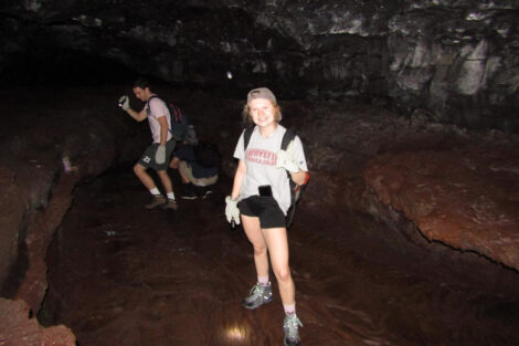 A young woman with light hair is standing in a cave. She's wearing a baseball cap and holding a backpack. A young man is crouched in the background and looking at the cave walls.