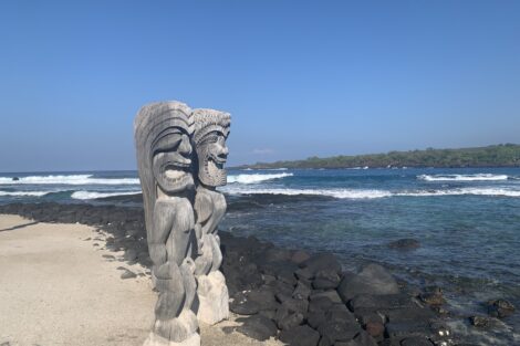 Two Hawaiian statues are overlooking a rocky beach.