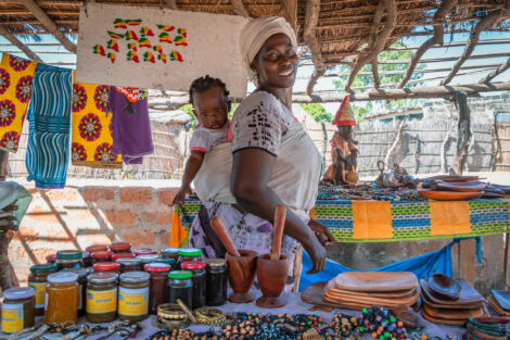 A Black woman is smiling directly at the camera with her baby strapped to her back. She's wearing white and has a headwrap. She's standing in a marketplace stall. There's a variety of spices and cups in front of her.