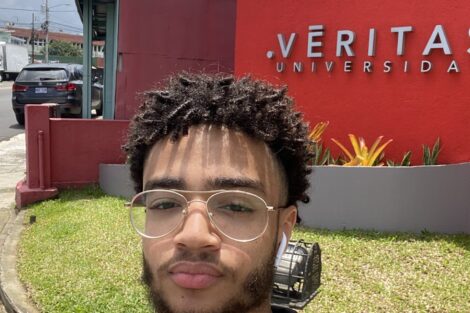 Young Afro-Latino student with curly hair and glasses stands in from of the Veritas University campus sign.