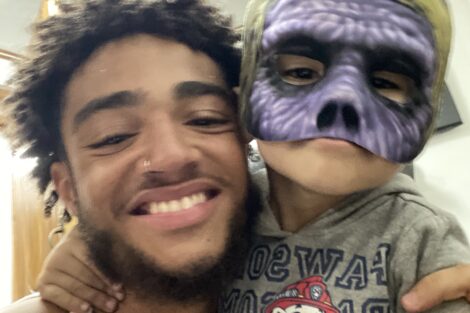 Young Afro-Latino student is smiling while hugging a young child wearing a costume mask.