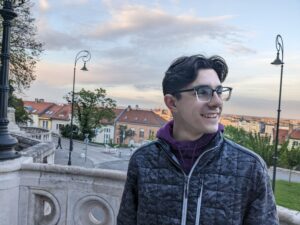 A young man with short brown hair and glasses looks to the side and smiles. He's standing on a marble balcony overlooking a suburban neighborhood in Bonn, Germany on a sunny day.