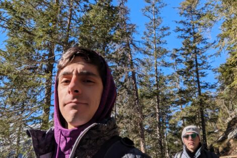 Two men wearing coats walking in a forest with tall trees.