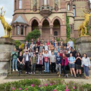 Large group of people standing on the steps outside a stone building. Bushes and large elk statues are on either side of the steps.